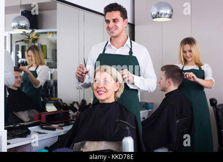 Lächelnd dunkelhaarigen Mann tun Frisur für reife blonde Frau im Friseursalon. selektive Fokus auf Frau Stockfoto