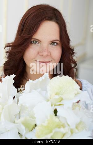 Porträt der schönen Frau mit Glücklich lächelnde Gesicht und braunem Haar in reinen weißen Kleid und Schmuck holding Blumenstrauß aus den weißen Blüten gekleidet auf Weiß Stockfoto