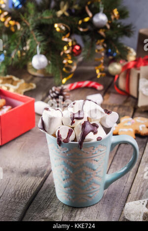 Traditionelle heiße Schokolade mit Marshmallows und Schokolade auf dem alten rustikalen Holztisch mit Weihnachtsbaum, Dekor, Geschenkboxen, Cookies und Gewürzen. Stockfoto