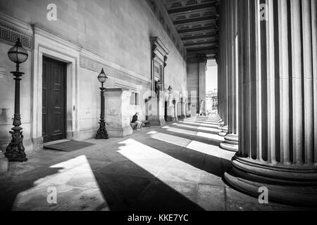 Licht und Schatten spielen auf der Säulen und die Architektur von St. George's Hall in lverpool Lime Street, North West England, mit einem Mann auf der integrierten Bank am oberen Ende der Stufen sitzend, auf einem schönen Sommer sonnigen Tag.. Stockfoto