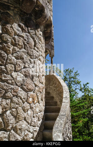 Schmale äußere Wendeltreppe, die sich um alte Turm aus natürlichen groben beige Steine gehen. Unscharfe blauer Himmel und grünen Park oder Wald Hintergrund. Stockfoto