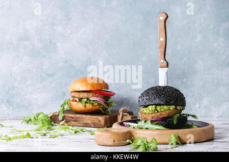 Satz von hausgemachten Burger in schwarze und weiße Brötchen mit Avocado, Tomaten, Rucola, Käse, Zwiebel auf Holz das Brett über weiße Holzbrett Tabelle Stockfoto