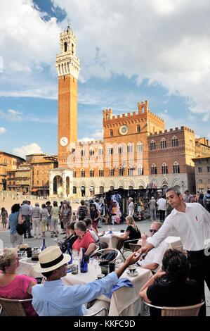 Touristen in Café am Piazza del Campo, dem zentralen Platz der Stadt Siena, Toskana, Italien. Torre del Mangia Turm erhebt sich hinter Stockfoto