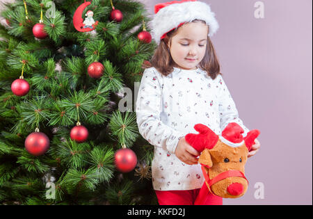 Kleines Mädchen, das zu Hause neben dem Weihnachtsbaum mit einem Plüschtier spielt Stockfoto