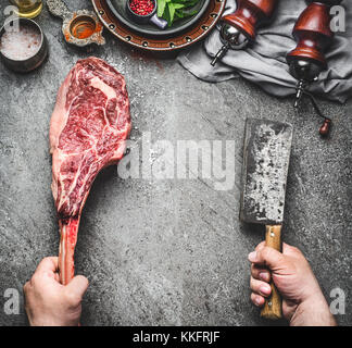 Männliche Hände der Metzger oder Koch Holding tomahawk Rindersteak und Fleisch Cleaver auf dunklen rustikalen Küche Tisch Hintergrund mit Zutaten zum Kochen und condi Stockfoto