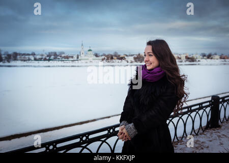 Junge Frau mit schönen langen Haaren in lila Schal steht in der Nähe des Snowy River an der Stadtpromenade und Wegsehen. kalter Tag, bewölkter Himmel Stockfoto