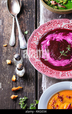 Sortiment von Creme Suppen aus Karotte, rote Bete und grüne Erbsen in verschiedenen Platte. Über alten Holztisch mit Vintage Löffel, Kräuter und Croutons. Rost Stockfoto