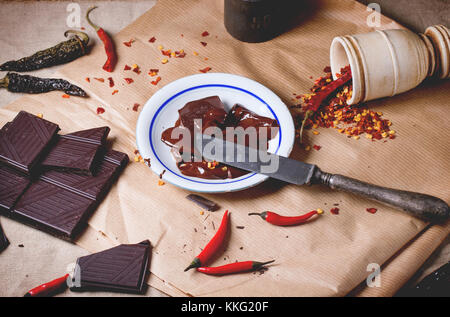 Häckseln, dunkle Schokolade mit frischen und trockenen red hot chili peppers auf Backpapier mit Teller von Heiß geschmolzene Schokolade und vintage Messer. Stockfoto