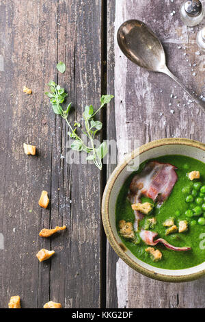 Cremesuppe von grüne Erbsen in Keramik Schüssel mit gebratenem Speck und Croutons. Über alten Holztisch mit Vintage Löffel, Salz und Kräutern. top anzeigen. Rus Stockfoto