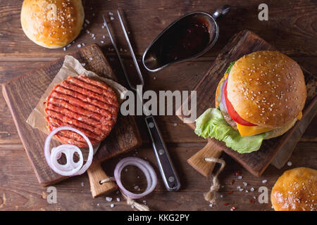 Frische, hausgemachte Burger auf wenig Schneidbrett und rohen Schnitzel und in Scheiben geschnittene Zwiebel, serviert mit Ketchup Soße und Fleisch Gabel über dunklen rustikalen Holztisch. Stockfoto