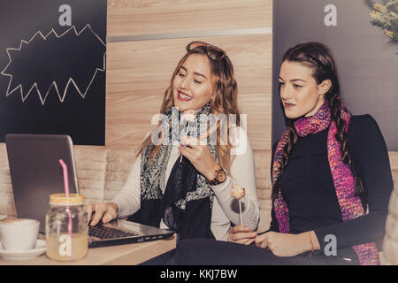 Zwei glückliche Mädchen mit Kuchen mit Laptop und trinken Kaffee im Cafe. Freundschaft und Technologie Stockfoto