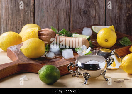 Haufen der ganze und aufgeschnittene Zitronen, Limetten und Minze mit Zitrusfrüchten Reibahle, Zucker und Eiswürfeln auf kleinen hölzernen Schneidebrett über Holz- Hintergrund. Rustikal Stockfoto