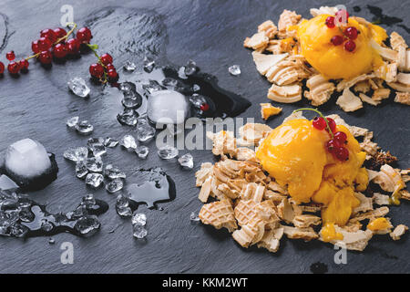 Mango sorbet Eis, serviert auf Wafer Krümel mit roten Johannisbeeren, Beeren und Eiswürfel über schwarzem Schiefer Stockfoto