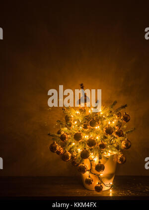 Kleine Weihnachtsbaum mit luxuriösen Spielereien und warme Beleuchtung, in einem weißen Kunststoff Blumentopf, mit dem dunklen Hintergrund. Stockfoto