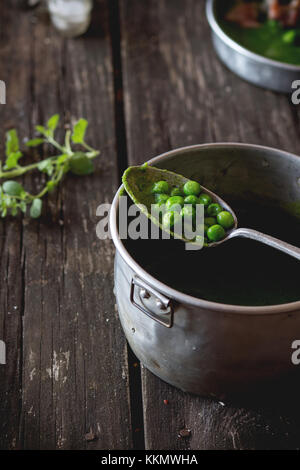 Cremesuppe von grüne Erbsen in Aluminium Pfanne mit Vintage Löffel füllen ganze Erbsen. Über alten Holztisch. dunklen rustikalen Stil, Tageslicht. Stockfoto