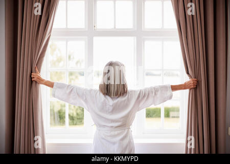 Ältere Frau Eröffnung Schlafzimmer Gardinen und Blick Aus Fenster Stockfoto