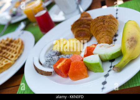 Leckeren, gesunden Frühstück im Freien an einem hölzernen Tisch mit Blick auf das Meer im Hintergrund. Das Frühstück besteht aus frischem Obst und Waffeln mit Pfannkuchen. Stockfoto