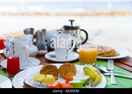 Leckeren, gesunden Frühstück im Freien an einem hölzernen Tisch mit Blick auf das Meer im Hintergrund. Das Frühstück besteht aus frischem Obst und Waffeln mit Pfannkuchen. Stockfoto