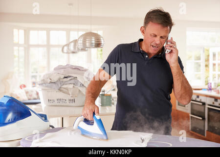 Im mittleren Alter Mann per Telefon abgelenkt beim Bügeln Stockfoto