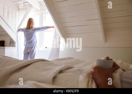 Frau öffnet die Vorhänge und schaut aus dem Fenster, als Mann liegt im Bett Stockfoto