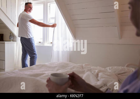Man öffnet die Vorhänge und schaut aus dem Fenster, wie Frau liegt im Bett Stockfoto
