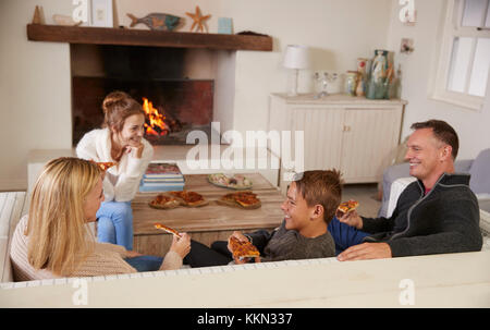 Familie sitzt auf einem Sofa in der Lounge am offenen Feuer essen Pizza Stockfoto