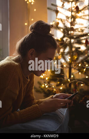 Frau mit ihrem Mobiltelefon in der Nähe von einem Weihnachtsbaum zu Hause Stockfoto