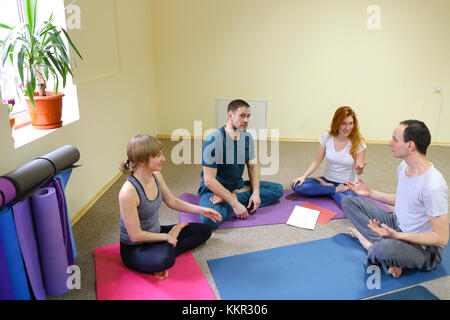 Zwei junge Frauen und zwei Männer sitzen im Fitness Studio im Erdgeschoss auf Yogamatten, Menschen mit Begeisterung Sprechen und Lachen. Freunde gekleidet in comforta Stockfoto