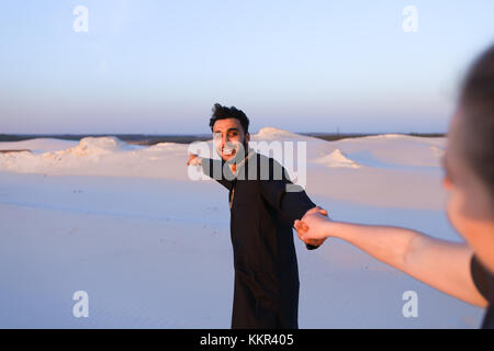 Junge muslimische Mann führt süßes Mädchen von Hand, Chatten mit junge Frau und lächelnd, das gilt auch für männliche. Liebhaber durch weite sandige Wüste am Fuß Stockfoto