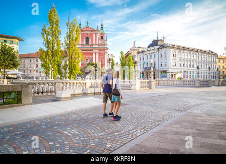 Ein paar Touristen, die in der Altstadt von Ljubljiana spazieren, mit der ikonischen Kirche der Franziskaner Verkündigung im Hintergrund. Ljubljiana, Osrednjeslovenska, Slowenien. Stockfoto