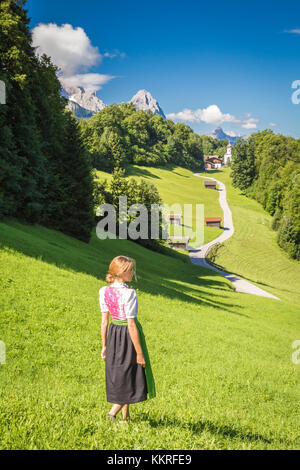 Ein Mädchen in typisch bayerischer Kleidung, das vor dem Dorf Wamberg spaziert, mit Zugspitze und Waxenstein im Hintergrund. Garmisch-Partenkirchen, Bayern, Deutschland. Stockfoto