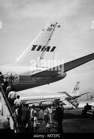 Im Jahr 1960 stiegen Passagiere am Flughafen Don Muang (DMK) in Bangkok, Thailand, in ein SAS DC-8-Flugzeug ein. Dieses Bild fängt die geschäftige Atmosphäre des Flughafens ein, während sich Reisende auf ihren Flug vorbereiteten. Dies markiert einen wichtigen Moment in der Luftfahrtgeschichte, als sich die Flugzeuge immer weiter verbreiteten. Stockfoto