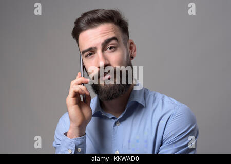Lustige Augenbraue verwirrten Gesichtsausdruck des Jungen Geschäftsmann am Telefon zu sprechen an Kamera auf grauen Hintergrund. Stockfoto