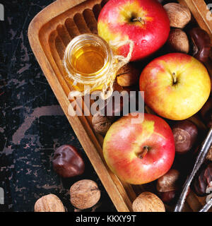 Glas Glas Honig mit Äpfel, Kastanien und Walnüsse in der hölzernen Platte über schwarzen Holztisch. top anzeigen. Bild. Stockfoto