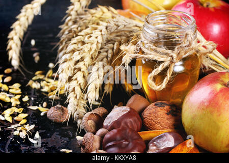 Glas Glas Honig mit Äpfeln, Ähren, Kastanien und Nüssen über schwarzen Holztisch Stockfoto