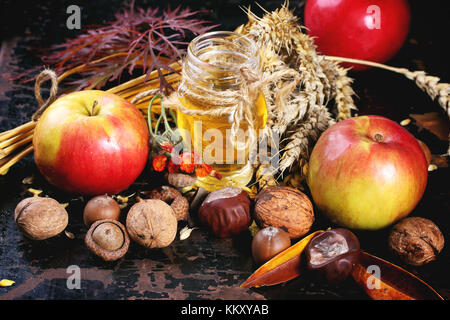 Glas Glas Honig mit Äpfeln, Ähren, Kastanien und Nüssen über schwarzen Holztisch Stockfoto