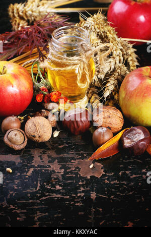 Glas Glas Honig mit Äpfeln, Ähren, Kastanien und Nüssen über schwarzen Holztisch Stockfoto