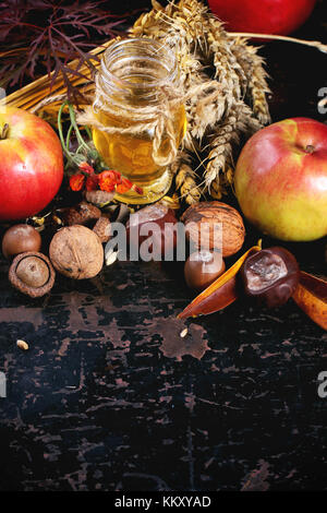 Glas Glas Honig mit Äpfeln, Ähren, Kastanien und Nüssen über schwarzen Holztisch. siehe Serie Stockfoto