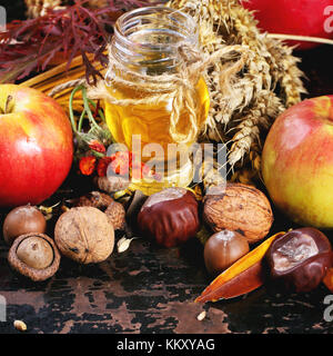 Glas Glas Honig mit Äpfeln, Ähren, Kastanien und Nüssen über schwarzen Holztisch. square Bild mit selektiven Fokus Stockfoto