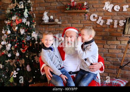 Schönen Jungen der gleichen Erscheinungsbild sitzen auf dem Schoß Santa Claus, der sitzt große und weiche Sessel Flüstern im Ohr gehegten Wünsche für das neue Jahr Gemütliche geräumige Zimmer Stockfoto