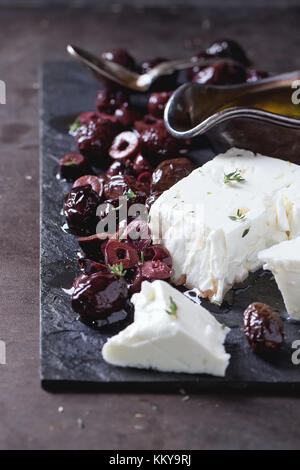 Ganze und aufgeschnittene schwarze Oliven und Feta Käse mit Olivenöl auf schwarzem Schiefer auf dunklem Hintergrund. Stockfoto