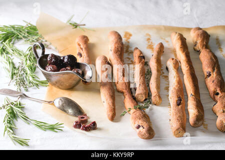 Ganze und aufgeschnittene schwarze Oliven in vintage Metallschale mit rasemary Kräuter und hausgemachte grissini Brot klebt auf weißen Tischtuch in der Sonne Licht. Stockfoto