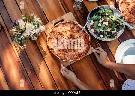Blick von oben auf die Frau Einstellung Pizza und Salat auf hölzernen Tisch für die einweihungsparty. Weibliche Vorbereitung einen Esstisch im Freien mit Speisen zum Abendessen. Stockfoto