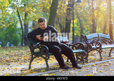 Im mittleren Alter Mann sitzt in einem Park mit einem Telefon Seite Portrait von glücklichen Mann sitzt auf einer Parkbank mit Handy Stockfoto