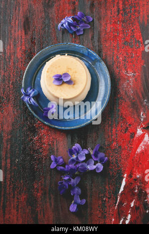 Ansicht von oben auf die Platte mit Karamell Pannacotta mit violetten Blüten über den roten Hintergrund Holz serviert. Stockfoto