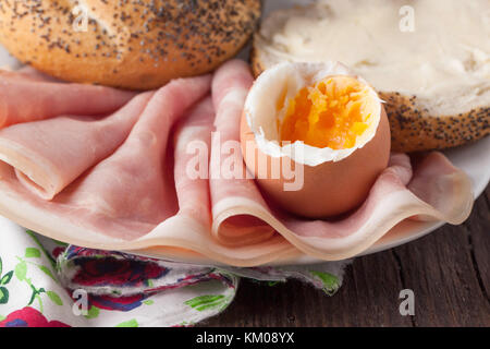 geschälte, gekochtes Ei, Textil, Brot mit Mohn und Butter, Schinken auf weißen Keramikplatte auf Holztisch Stockfoto