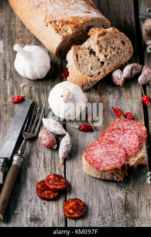 Sandwich mit slised Salami mit anderen Würstchen serviert, frisches Brot, Knoblauch, red hot chili peppers und vintage Besteck auf alten Holztisch. siehe Serie Stockfoto