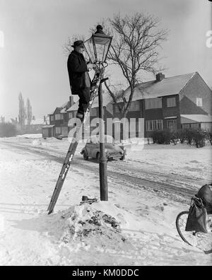 Bild am 22. Januar 1963 genommen, der Staffordshire Dorf Pelsall, zeigt einen Mann an der Spitze von einer Leiter, die Beleuchtung der gasbetriebenen Straßenlaternen. Ein Winter mit Schnee auf dem Boden und einen Morris Monor Auto im Hintergrund. Stockfoto