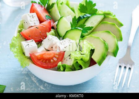 Grüner Salat mit Avocado in Scheiben, Tomaten, Gurken und griechischer Schafskäse in Schüssel über blauen Tabelle. gesunde Ernährung vegan vegetarisch Sommer Gemüse Sal Stockfoto