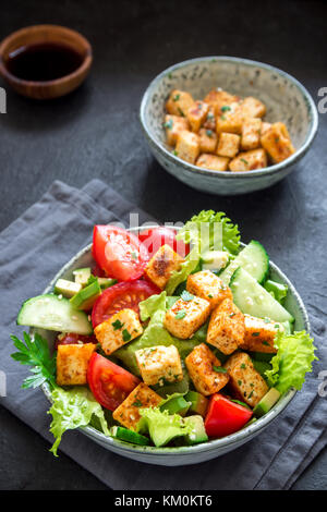 Gebratenem tofu Salat mit Gurken, Tomaten, Avocado und Sesamsamen. Hausgemachte asiatische Gemüse und Tofu Salat in Keramik Schüssel auf schwarzen Stein Hintergrund Stockfoto